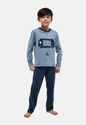 Garçon debout pieds nus portant une chemise à manches longues bleue avec une manette de jeu et le texte « PROCHAIN JOUEUR », ainsi qu'un pantalon bleu foncé sur un fond uni.