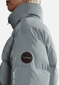 Lys grå pufferjakke med høj krave, der har et cirkulært logo-patch på overarmen og en slank, glat tekstur.