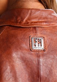 Brun läderjacka med en texturerad yta, försedd med en rektangulär metall-logotyp som är märkt "Freaky Nation" på baksidan.