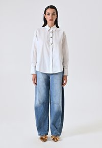 Camicia bianca a maniche lunghe con colletto a punta e bottoni dorati, abbinata a jeans a gamba larga blu chiaro e scarpe decorate con fiocchi dorati.