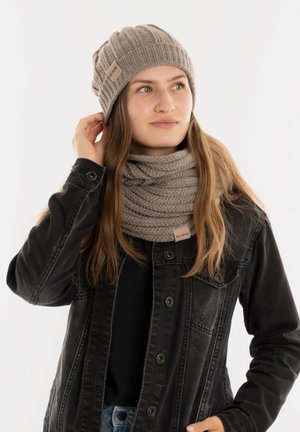 Beige gebreide beanie en bijpassende chunky sjaal op een model. De sjaal heeft een ribbelstructuur. Beide accessoires hebben kleine merklabels.