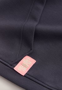 Gros plan d’une couture de tissu foncé avec une étiquette rectangulaire rose cousue, comportant un texte en creux.