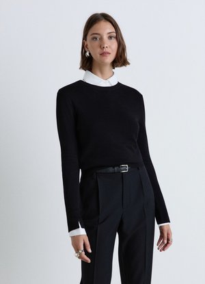 Donna con capelli corti castani che indossa un maglione nero sopra una camicia bianca con colletto e pantaloni neri a vita alta con cintura, in piedi davanti a uno sfondo semplice.
