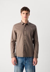 Camicia marrone a maniche lunghe con bottoni, righe verticali rosse, bottoni neri e colletto puntato, abbinata a jeans azzurri chiari.