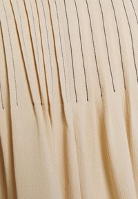 Tessuto beige plissettato con sottili linee verticali, caratterizzato da cuciture scure a contrasto. Texture morbida, materiale leggero, drappeggio elegante.