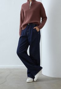 Gerippter, brauner Pullover mit Reißverschluss am Hals und langen Ärmeln, kombiniert mit locker sitzenden, dunkelblauen Jeans und weißen Sneakers, stehend vor einer hellen Wand.