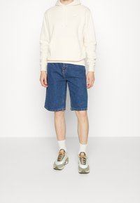 Ljusbeige hoodie med en framficka och logotyp, kombinerad med knä-långa blå denimshorts och flerfärgade sneakers, stående mot en vit bakgrund.