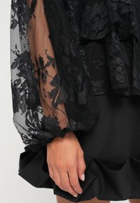 Sort kjole med gennemsigtige blonde lange ærmer med blomstermønstre, suppleret af en flæse detaljeret sort skørtekant. Teksturdetaljer er synlige.