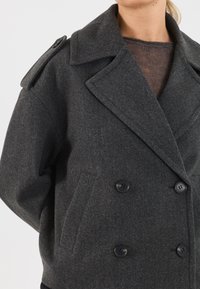 Donna che indossa un cappotto di lana grigio scuro a doppio petto con ampi revers e una tracolla, sopra un top in maglia marrone, su uno sfondo bianco.