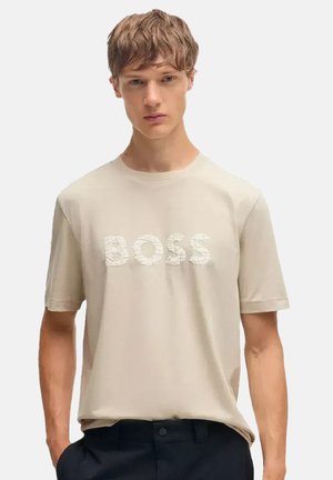 Młody mężczyzna o krótkich, jasnobrązowych włosach, ubrany w beżowy t-shirt z tekstem o fakturze ""BOSS"" i ciemne spodnie na jednolitym tle.