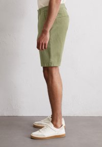 Khaki green shorts made of lightweight fabric, featuring a flat front, side pockets, and back pocket. Paired with white sneakers and gum soles.