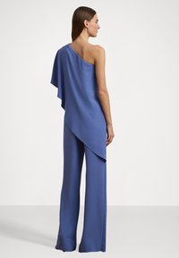 Asymmetrisk blå jumpsuit i let materiale, med én skulder, vide ben og en flydende draperet detalje bagpå.