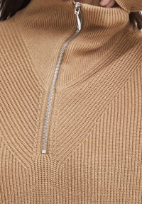 Pullover in maglia color nocciola con collo alto e chiusura a zip. Motivo testurizzato con coste verticali e un design a forma di V sul davanti.