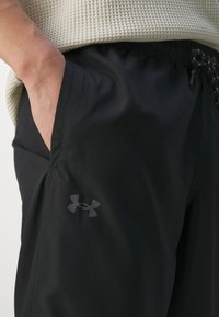 Fekete sportnadrág zsebében a kéz, amelyen az Under Armour logó található, bézs textúrájú toppal párosítva.