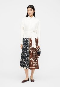 Γυναίκα όρθια, φορώντας λευκή μπλούζα με ζωνάκι, μίντι φούστα με μαύρα και καφέ λουλουδένια σχέδια και σκούρα καφέ φλατ παπούτσια, σε λευκό φόντο.