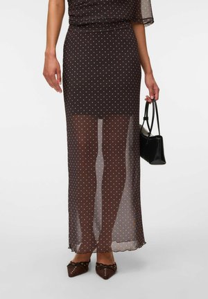 Femme portant une jupe marron transparente à pois par-dessus une jupe plus courte, tenant un petit sac à main noir et portant des chaussures pointues marron.