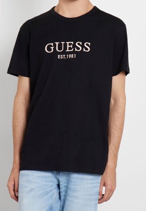 Uomo con t-shirt nera GUESS con scritta "EST. 1981" abbinata a jeans azzurri chiari su sfondo semplice.