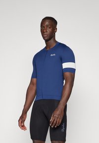 Maillot de ciclismo azul marino con un diseño ajustado, mangas cortas, tejido texturizado y una franja blanca en el brazo izquierdo. Pantalones cortos de ciclismo negros con logo.