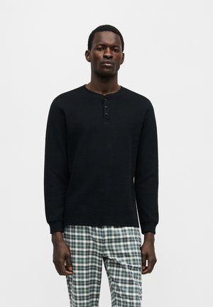 Mężczyzna stojący, ubrany w czarną koszulkę henley z długim rękawem oraz zielone i białe spodnie w kratę na jednolitym jasnym tle.