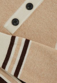 Dziany sweter w beżowej wełnie z dwoma czarnymi guzikami. Żebrowane mankiety mają pasek w kolorze kremowym i brązowym. Miękka, teksturowana tkanina.