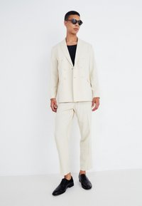 Blazer de color beige claro, de doble botonadura y con solapas de muesca, combinado con pantalones a juego. Unos zapatos de cuero negros y unas gafas de sol redondas completan el look.