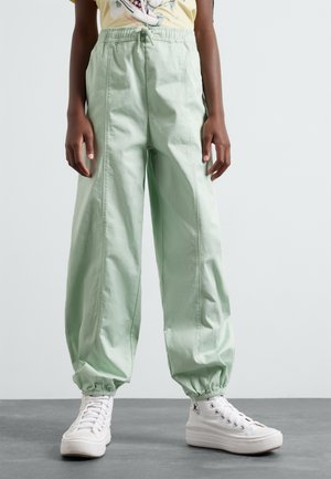 Pantalones sueltos de color verde claro con una cinturilla elástica y puños. Combinados con zapatillas altas blancas sobre una superficie gris.