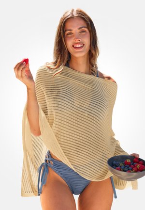 Femme en bikini bleu avec un châle beige tenant un bol de baies mélangées et une seule fraise, souriant à la caméra.