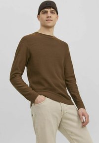 Pull marron à côtes, à manches longues, avec un col rond, associé à un jean beige clair ; le mannequin a les mains dans les poches et porte un bonnet noir.