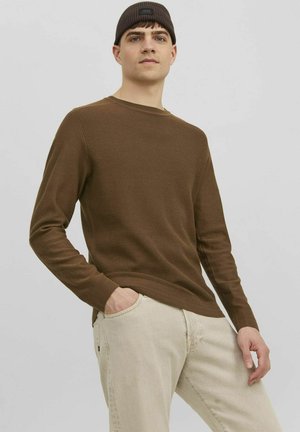 Pull marron à côtes, à manches longues, avec un col rond, associé à un jean beige clair ; le mannequin a les mains dans les poches et porte un bonnet noir.