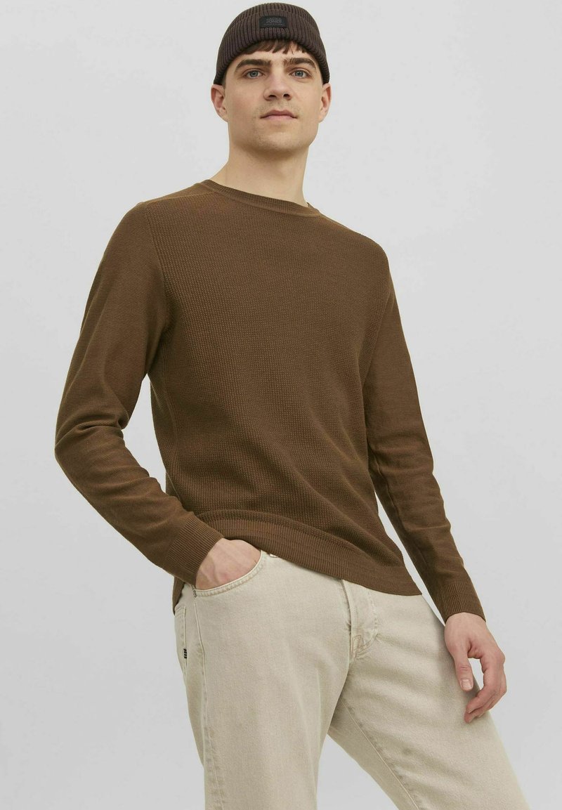 Pull marron à côtes, à manches longues, avec un col rond, associé à un jean beige clair ; le mannequin a les mains dans les poches et porte un bonnet noir.