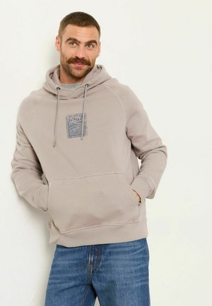 Mann mit Schnurrbart, der einen beigen Hoodie und blaue Jeans trägt, entspannt mit den Händen in den Taschen vor einem hellen Hintergrund steht.