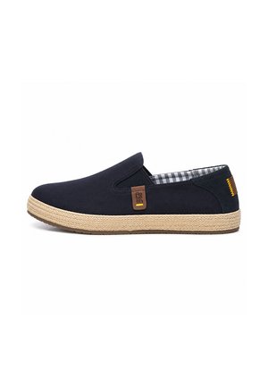 Zapato azul marino tipo mocasín con suela beige tejida, forro interior a cuadros, paneles laterales elásticos y etiqueta de logo marrón en el lateral exterior.
