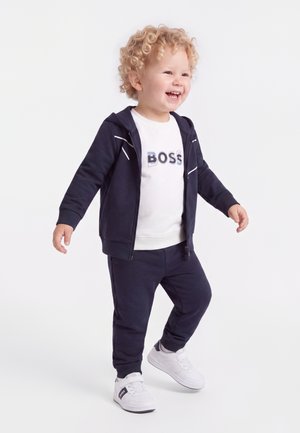 Tout-petit aux cheveux blonds bouclés sourit, portant un survêtement bleu marine, un t-shirt blanc et des baskets blanches sur un fond clair uni.