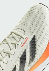 Jasnoszare buty sportowe z teksturowaną siateczkową cholewką, czarnymi akcentami, pomarańczową gradientową podeszwą i kontrastowymi sznurowadłami. Charakteryzują się smukłym wzornictwem.