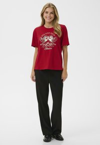 Raudona grafika marškiniai su sidabriniu erelio dizainu ir tekstu "Skristi per aukštai" ir "Laisvė." Derinama su juodais tiesiais auditorijos kelnėmis ir juodais batais.