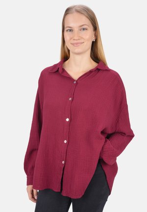 Burgundy button-up shirt in textured fabric, featuring a collar, long sleeves, and side slits, paired with black pants.