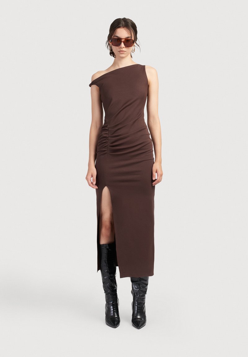 Hnedé šaty na jedno rameno s bočným riasením a rozparkom až po stehno, vyrobené z jemnej látky. V kombinácii s čiernymi vysokými čižmami a slnečnými okuliarmi.