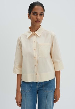 Vrouw die een crèmekleurig overhemd met knopen en driekwart mouwen draagt, gecombineerd met een blauwe spijkerbroek met voorzakken, staand tegen een effen achtergrond.