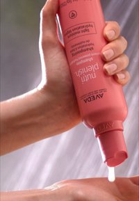 Bouteille rose de produit capillaire Aveda Fruit Finishing tenue au-dessus d'une main. Le produit a une texture lisse, et une petite quantité s'écoule dans la paume.