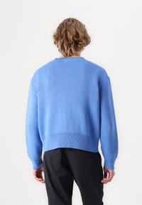 Pulover tricotat albastru cu guler rotund și manșete cu nervuri, prezentând un tiv ușor scurtat și un finisaj texturat. Se poartă pantaloni negri.