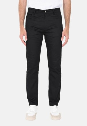 Uomo che indossa pantaloni neri slim fit e sneakers bianche, in piedi davanti a uno sfondo chiaro e uniforme.