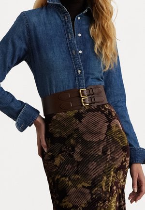 Kobieta ubrana w ciemnoniebieską koszulę dżinsową zapinaną na guziki, wpuszczoną w wysokociśnioną kwiatową spódnicę z szerokim brązowym paskiem z podwójną klamrą.