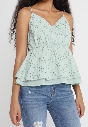 Femme portant un haut sans manches en dentelle ajourée vert clair avec un ourlet à basque et un jean bleu déchiré, debout devant un fond uni.