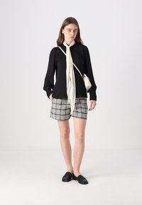 Svart langermet bluse med blonde detaljer, hvit halsduk, grå rutete shorts og svarte loafers; minimalistisk hvit håndveske.