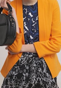Orange blazer with structured design, paired with a patterned blue and white top and black marbled skirt. A small black handbag is held.