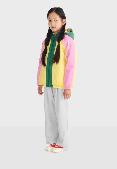 Jong meisje met lang gevlochten haar, gekleed in een colorblock-jas met groene, gele en roze delen, grijze broek en rode schoenen met witte zolen.