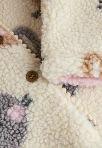 Fleecejacke mit einer weichen, flauschigen Textur in Creme, ausgestattet mit braunen Knöpfen und einem Muster aus pastellfarbenen Formen auf dem Stoff.
