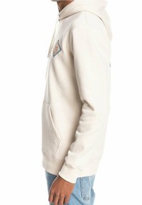 Světle béžová mikina s přední kapsou, nastavovací kapucí na šňůrku a detailem nášivky na hrudi. Hladký bavlněný materiál.