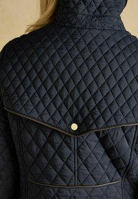 Námornícky prešívaný kabát s vysokým golierom, diamantovým prešívaním a zlatým cvokom na zadnej strane. Diskrétny hnedý lem.