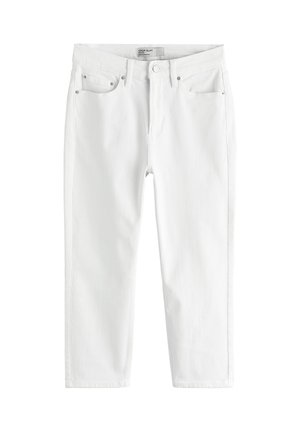 Jeune femme portant un top rayé à col halter et un pantalon blanc, appuyée contre un mur blanc près d'une piscine avec l'océan en arrière-plan.
